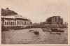 The front of a postcard showing small fishing boats along the edge of the water and two large buildings behind them on land. 