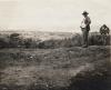 Humphrey Tracy standing on a hill looking at Port of Spain. He is wearing a suit and hat. There are trees and water in the background.