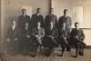 A photo of four International Drillers standing and six sitting in chairs on a boat deck. They are wearing dark suits and ties. They are in front of a wall that has five columns of rivets in it. 