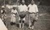 Two native boys standing beside Tracy and Hicks, who are wearing light-coloured clothing and are holding hats. 