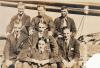 Seven men wearing ties and dark suits sitting in three rows (three, three, and one at the bottom). 