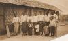 Une photo de sept femmes et quatre enfants se tenant devant un édifice en chaume. Les femmes portent des jupes et des chemisiers blancs. 