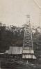 A photo of a covered oil rig. There is a group of men in front of it. There is casing in the foreground and trees in the background. 