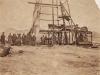 A photo of a group of men wearing work clothes stand at the base of a metal oil rig. There is a piece of cloth covering the left side of the rig. The ground is bare and there are hills in the background. 