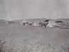 A photo of three white tents in the desert with two black tents behind. There are several people around the tents and one riding a donkey in the foreground. 