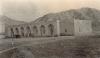 A photo of a bungalow with a flat roof and seven arches at its front. There are a group of people and a horse standing in front of it, and hills behind it. 