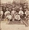 A photo of three patterned rugs on the ground. Five International Drillers are sitting in chairs and two are sitting on the ground. There are bushes behind them. 