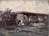 A photo of a thatched bungalow. There is a group of people outside the house, and two of the men are in uniform. There are trees on either side of the photo. 