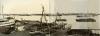 A photo of boats tied up to a dock. There are building materials and metal tracks on the dock. More boats float out in the water behind and there are buildings along the far bank of the water.