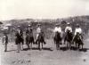 Six International Drillers on horseback. They are wearing pith helmets. A Burmese man stands to their left. There are trees and bushes in the background.