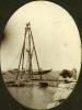 A photo of three men on a wooden tower wit four poles and ropes leading down to a dock. A boat with its mast down is anchored in the water behind them. There is a fourth man on the right beside a pile of objects wrapped in dark material.