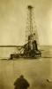 An oil derrick covered halfway up in metal siding. There is mechanical equipment on the right. The ground is flat dirt and there is a hill in the background. The photographer's shadow is at the front of the photo.