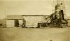 An oil derrick covered in metal siding. There is mechanical equipment on the right. The ground is flat dirt and there is a hill in the background.