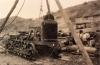 A photo of a partially assembled bulldozer beneath a wooden tripod. There is one man at the front of the bulldozer and four behind it. There are hills in the background. 