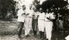 A photo of five men wearing white shirts stand together, holding small glasses of alcohol. There is a group of people behind them to the right.