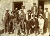 A photo of a group of International Drillers standing and sitting in front of a light stone building with an open doorway. They are wearing suits and vests.