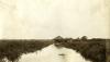 A thatched bungalow on stilts in the middle of a marsh. There is a strip of open water down the centre of the photo.