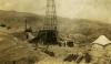 A metal oil rig. There are men standing around it and near a car. There are metal cylinders in the foreground and a tent on either side of the rig. There is a cylindrical steam boiler behind the rig. There are hills in the background.