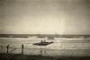 A photo of two men standing on a raft in shallow water and two on the beach. There are two ships anchored off-shore. 