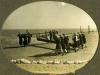 A photo of a group of men carrying a pipe. The pipe is attached to two beams and the men are standing at the front and back of the pipe and supporting the beam on their backs. The ground is flat and there is a line of white rocks in the foreground.