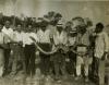 Nine men in light-coloured shirts. The five on the right are holding a snake stretched between them. There are trees in the background.