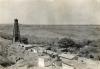 A single oil rig in an open plain with piles of timber and casing beside a few small buildings. 