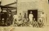 Six men standing and eight men sitting in front of a stone building with a set of wooden doors that are open.