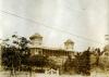 A photo of a building in behind trees and a gate. There are turrets on the corners of the building that are visible.