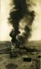 Thick dark smoke coming off of an oil well. There is a group of people off to the side and equipment in the foreground. There are hills in the background.