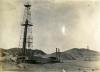 An oil rig with four legs and crossbeams for support. There are four lines attaching it to the ground and there is a covered building at its base. There are dirt hills and another rig in the background.