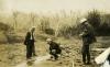 Four men looking down at a small water spring running through a bed of dirt and rocks. There are trees with no leave in the background.