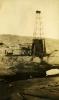 A metal oil rig. The base is covered in dark cloth. There is a roof over the wheels and belts beside the rig. There is casing on the ground in front of the rig and a bare hill in the background.