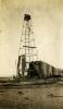 An oil derrick with four poles. The pole on the right is bent out of shape. There are three men on the top of the derrick and another climbing towards the, There is a group of men at the bottom of the rig, and part of the rig is covered in dark cloth.