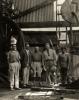 Group of five men in dirty clothing standing inside an oil rig covered in sheeting. There is a large wheel and belt behind them. 