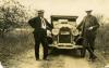 Webb and Burns standing on either side of a car parked on a dirt road. There are short trees on either side of them. The licence plate between the headlights reads: 1924. I.P.Co. STA.ELENA. 40".