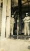 Two men wearing work clothes and hats standing inside a drilling rig. There is a large pipe and a wheel in behind them.