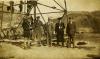 Five International Drillers standing at the base of a metal oil right, wearing dark suits and hats. There are men working on the lowest crossbeam. There are hills in the background.
