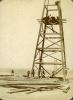 A wooden oil derrick with four legs. A group of men stand at its base surrounded by a pile of wooden planks. Three men are on the derrick and there are two ladders resting against the lowest support beam.