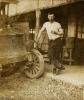 Ernest Kells wearing a white t-shirt and dark pants. He is resting his hand just above the wheel of a car and is standing in front of a building.