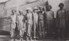 Seven men standing in front of a structure covered in metal siding. Eric Hussey stands in the middle with his arms around two men. He is wearing a pith helmet and coveralls.