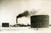 A photo of two large cylindrical storage tanks above ground, There are buildings and a smoke stack producing black smoke behind them. There is a railway track going across the foreground.