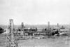 Three oil derricks stand in a field in front of a group of buildings. There are pipes connecting the buildings. At the bottom of the picture, "Woodward's Refinery" is written in white.