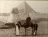 John Growder sitting on a camel in front of the Sphinx and a pyramid. There is a man holding the reins of the camel and another man at the back of the camel.