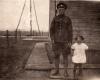 Arthur Brown standing next to Dorothy. He is wearing a military uniform and she is in a white dress. They are standing in front of a wooden building with wooden planks around it. There are derricks in the background.