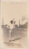 Gilbert Brown standing in front of a wire fence, holding a cigarette. He is wearing a white shirt and pants, with a boat hat. There are two oil derricks in the background.