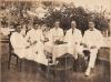 Five men sit around a wicker table covered with a white tablecloth. There is a bouquet of flowers in front of a frame on the table. The men are wearing white suits.